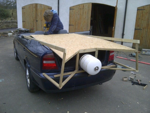 A boy cuts wood on top of a car with a Batman framework nailed on