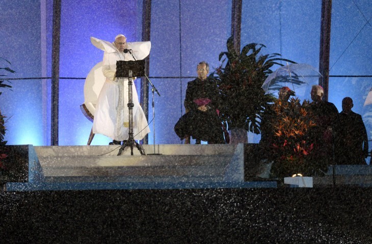 Photo of Pope Francis speaking on stage, wind whipping his robes