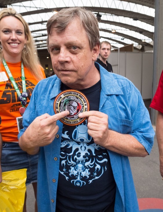 An older Mark Hamill holds up Star Wars decal for the camera at a fan convention