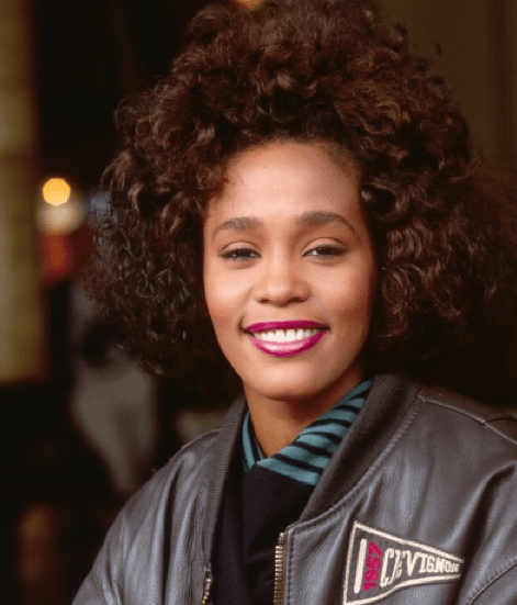 Photo of Whitney Houston, young and smiling warmly in a leather coat