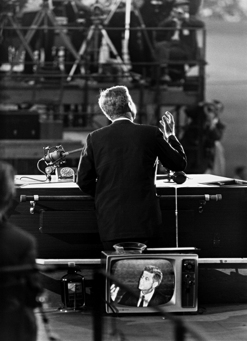 A photo of John F. Kennedy seen from behind as he speaks to the convention hall; a TV showing his image is in the foreground