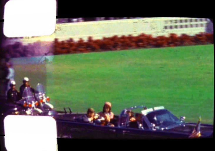 A Zapruder home movie frame showing JFK and Jackie Kennedy in the motorcade before a bright green lawn