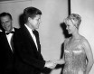 JFK and Ralph Bellamy wear tuxes; Dorothy Provine is a blonde wearing a flapper-style cap and beaded dress. She smiles as she shakes JFK's hand in some kind of function room.