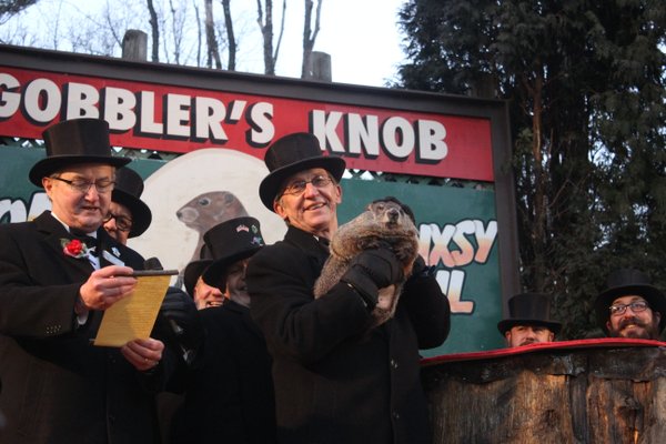Photo of Punxsutawney Phil being held in the air by a man in a top hat, in front of a sign reading GOBBLER'S KNOB