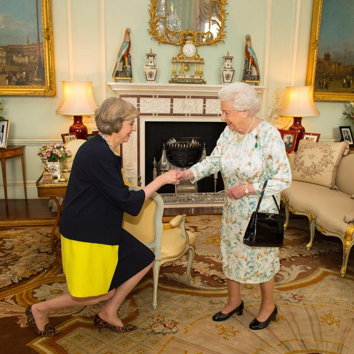 Theresa May in an odd blue-and-yellow skirt curtseys before a tiny Queen Elizabeth II in a formal dining room in Buckingham Palace