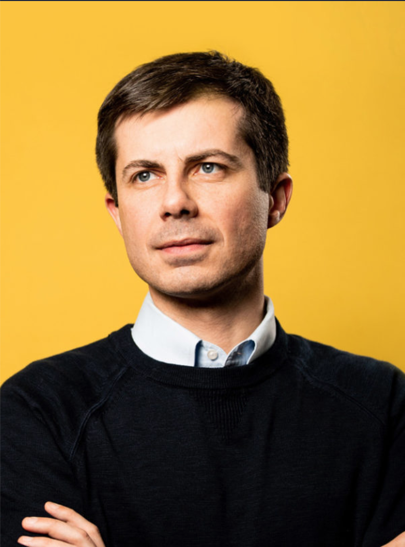 Pete Buttigieg wears a dark crew-neck sweater, with a button-down collar peeking over the top. He is clean-shaven and has a full head of dark hair.