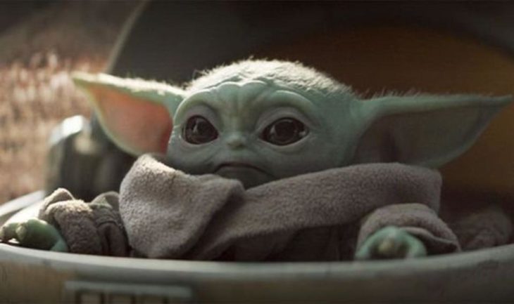 Baby Yoda has huge horizontal ears, wide black eyes, and a scrunched-up (but cute!) face as he sits in a clamshell-type pod