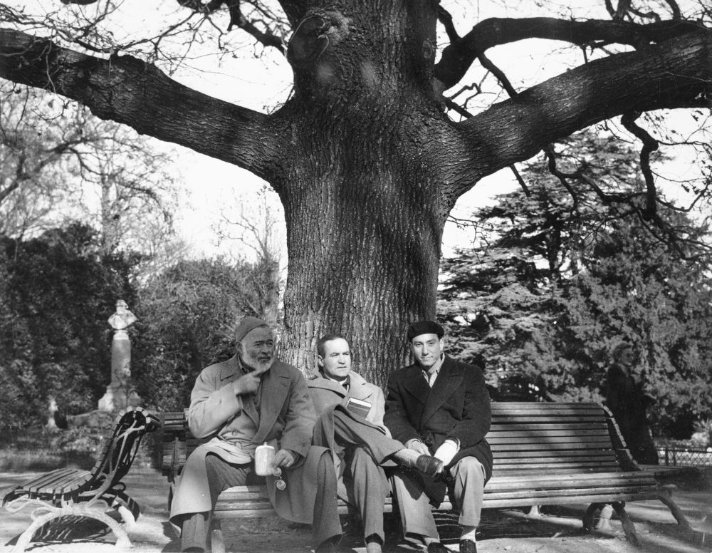 Ernest Hemingway looks middle-aged and jovial and holds a flask; all three men are bundled up for spring-like weather under a big old tree in what looks like a park
