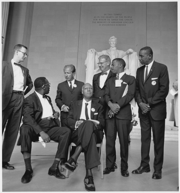 Seven youngish men in suits chat together at the Lincoln Memorial, in front of the famous statue of Abraham Lincoln