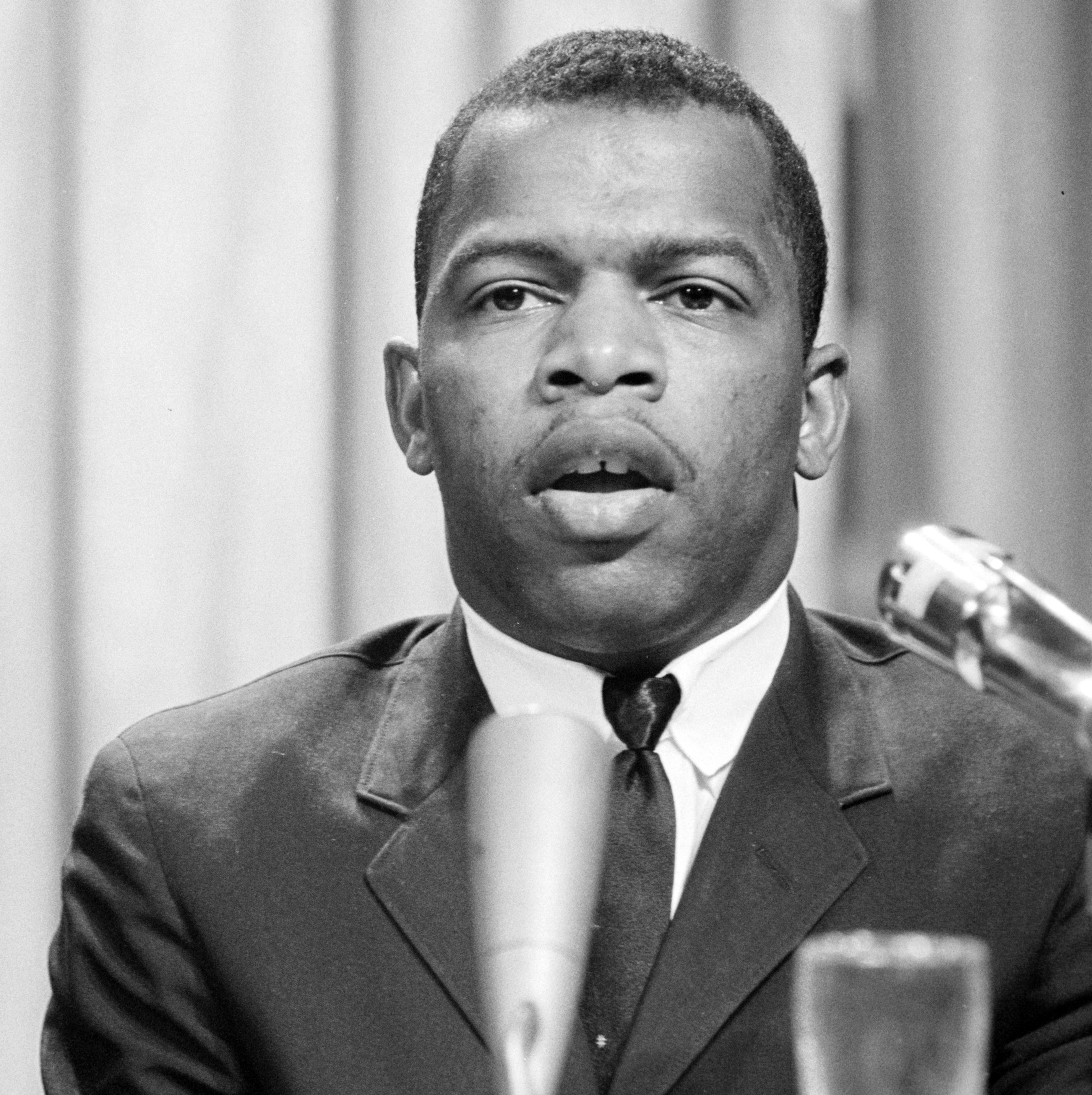 John Lewis, wearing a dark suit and tie, speaks behind a lectern in front of a rather bland gray backdrop