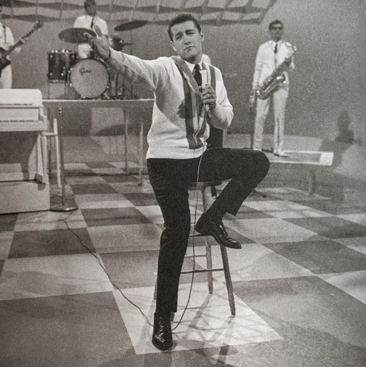 A young Alex Trebek sits on a high stool, leg cocked, holding a mic and wearing an old-fashioned sweater that was probably hip casual in those days. Behind him are musicians with a guitar, a drum kit, and a saxophone