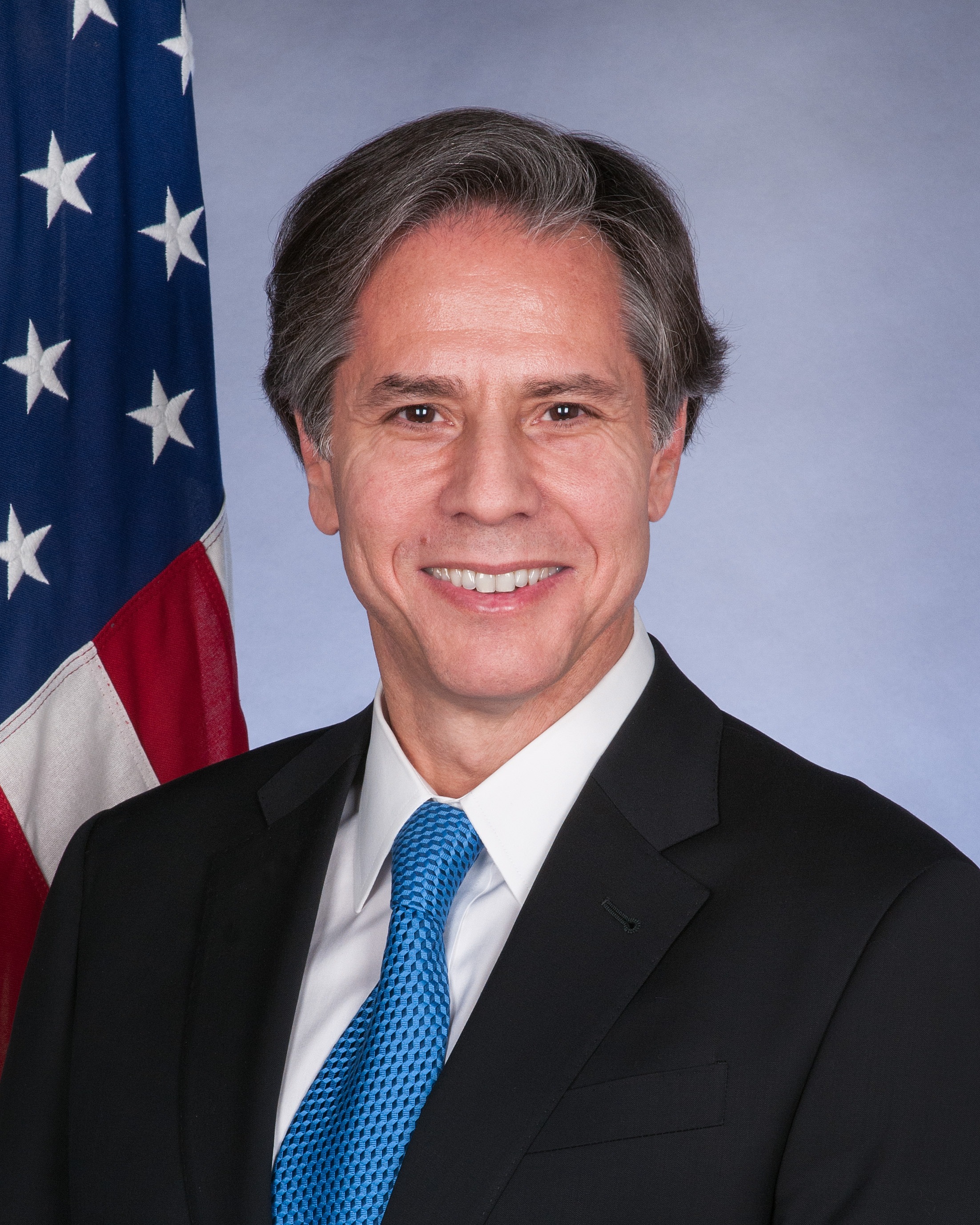 Antony Blinken poses in the classic government shot: blue suit and tie in front of an American flag