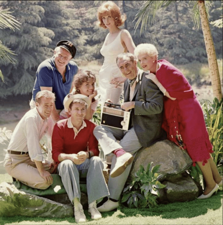 Seven people in casual dress sit and stand around a tree trunk on an (alleged) tropical island