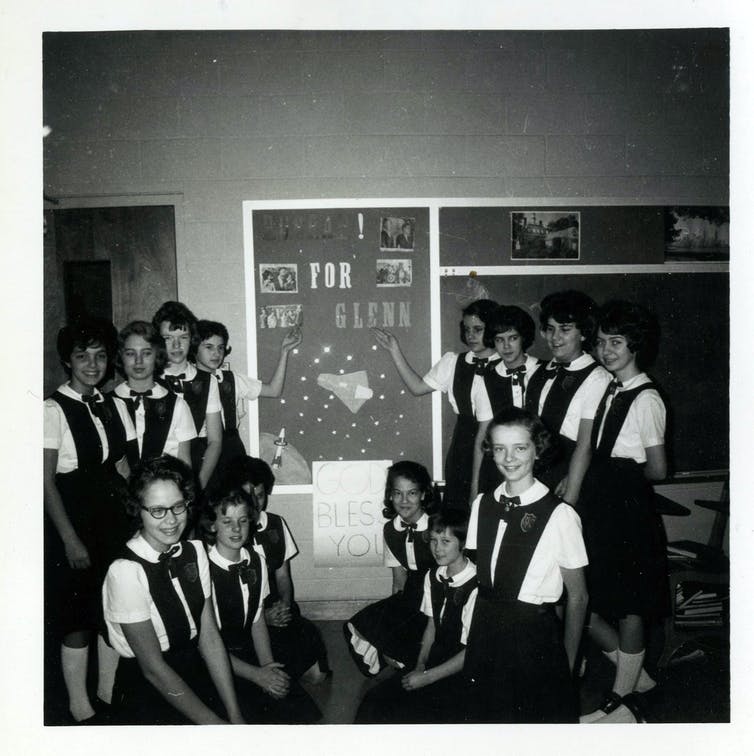 Black and white photo of uniformed schoolgirls in classroom gathered around a handmade poster cheering on John Glenn 