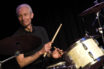 A smiling gray-haired Charlie Watts sits behind a drum kit, looking genteel as he plays
