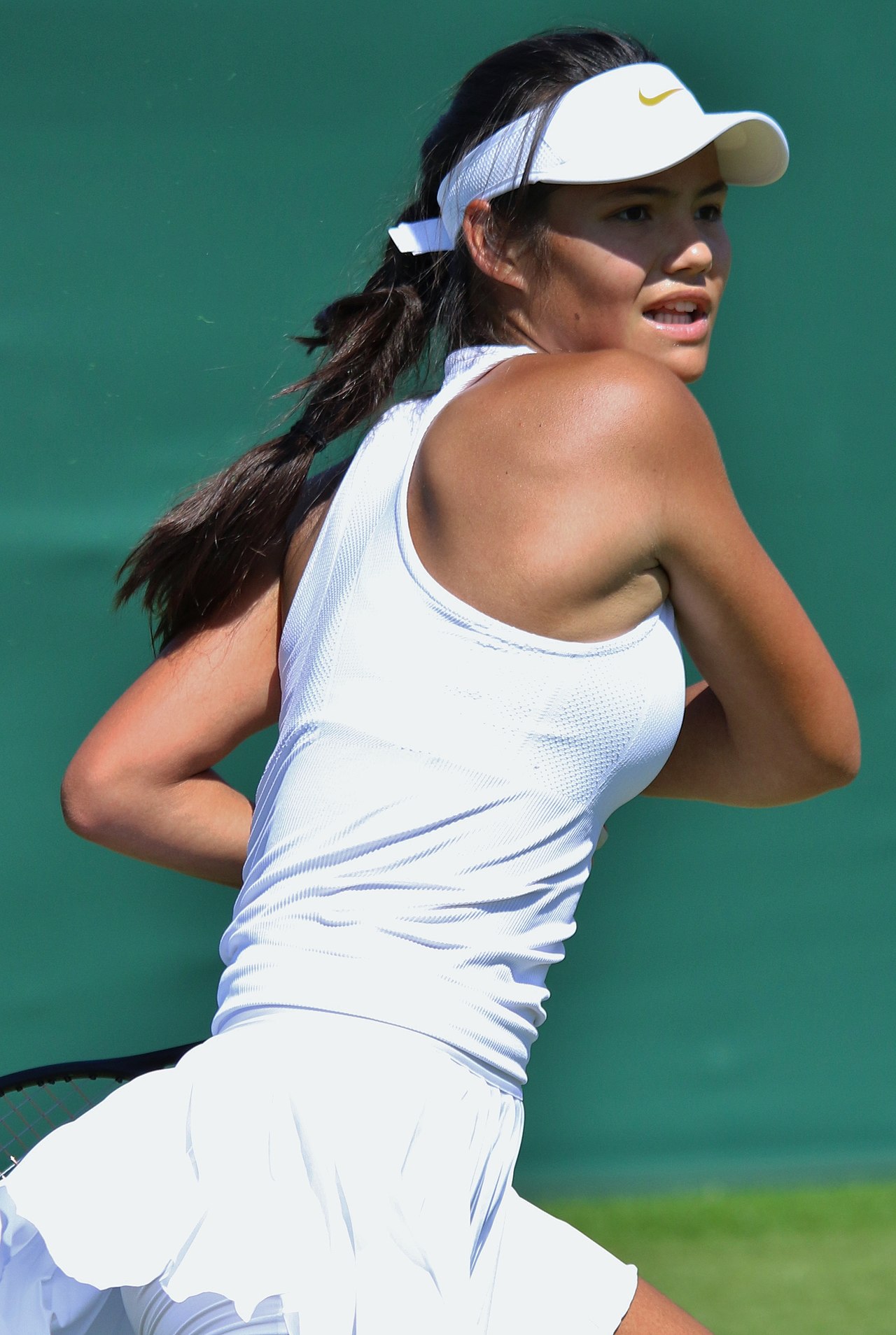 Emma Radacanu dashes cross-court for a shot. She wears a white tennis dress and a white visor, in accordance with the Wimbledon dress code