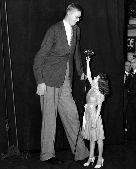 An astoundingly tall young man, Robert Wadlow towers over a 5-foot-tallish woman as they stand together at an old-time radio microphone