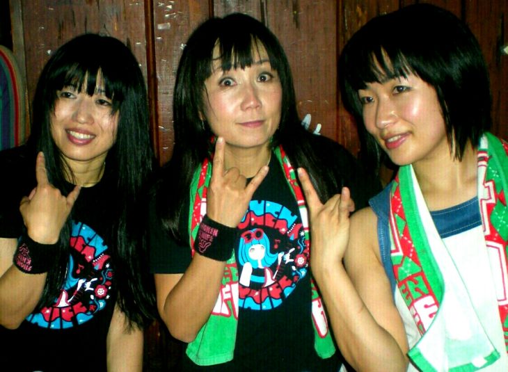 Three sweaty Japanese women smile as they hold up their hands in horns with their second and fifth fingers