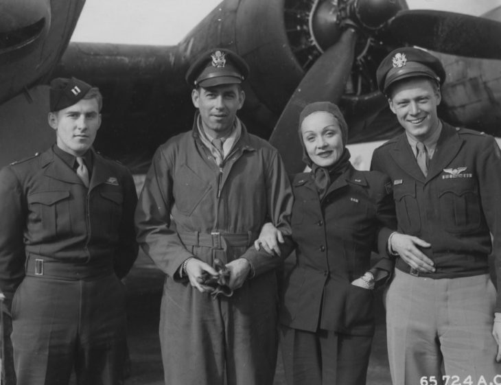 Marlene Dietrich, head wrapped in a scarf, poses with three pilots in front of a World War II airplane