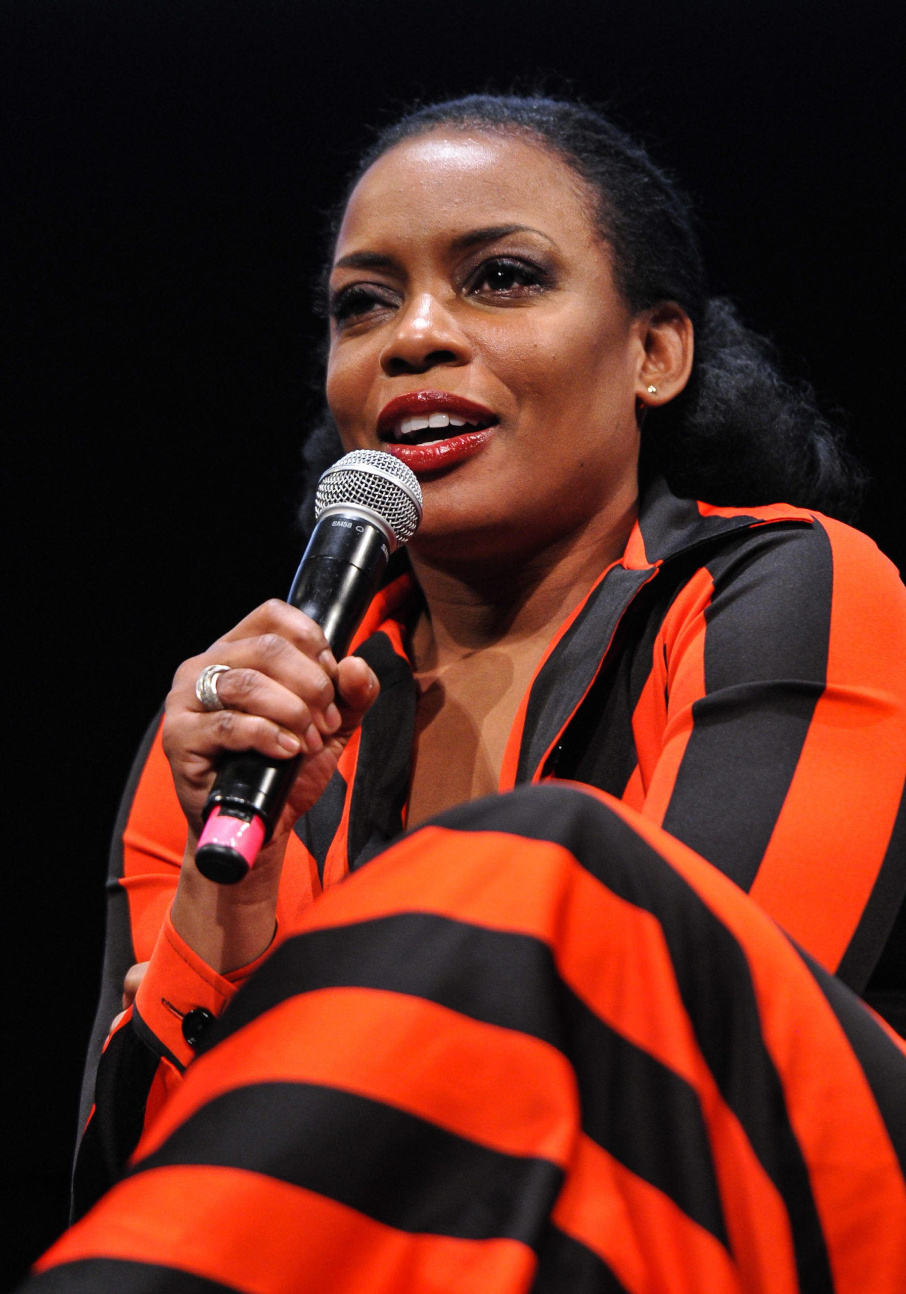 Aunjanue Ellis is a black woman in a striking striped gown, speaking into a microphone while sitting onstage
