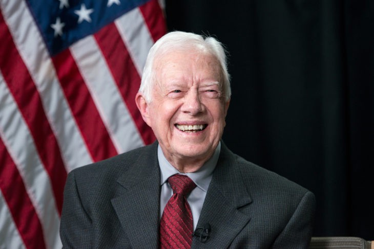 A gray-haired man smiles in front of an American flag