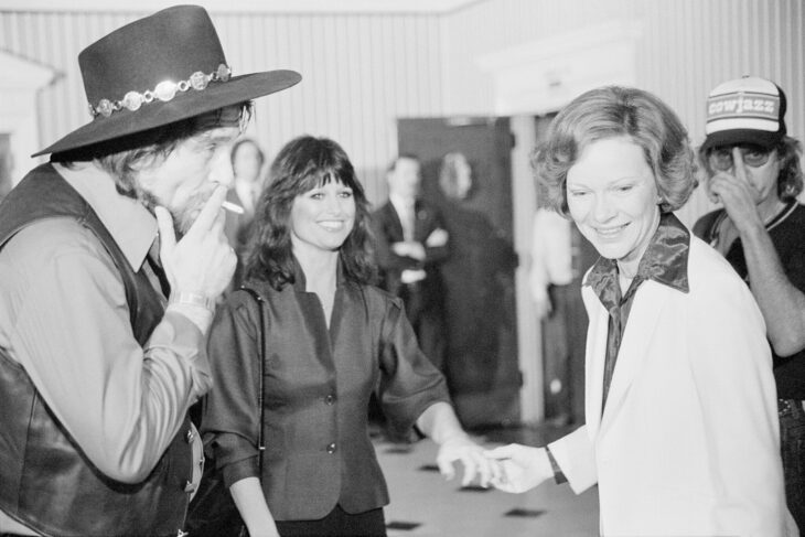 Rosalynn Carter smiles as she chats with Waylon Jennings (in a leather vest and cowboy hat and smoking a cigarette) and Jesse Colter in a fancy White House room. 