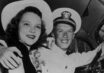 Two very young people, one in a naval officer's uniform, smile in a car.